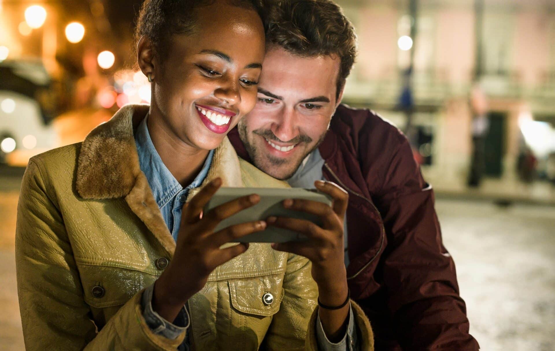 Portrait of happy young couple looking together at smartphone by night, Lisbon, Portugal